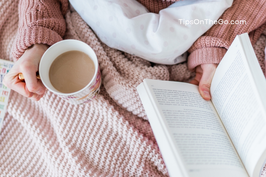 A person reading a book and drinking coffee on their bed.