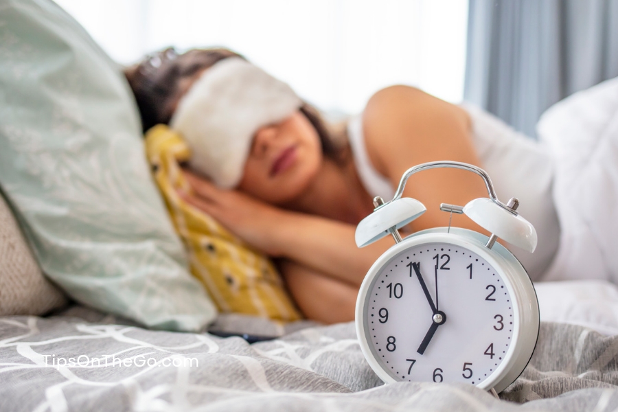 A woman wearing a blindfold is sleeping next to an alarm clock.